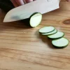 Mains utilisant un couteau de cuisine Sanakaza pour couper des rondelles fines de courgette sur une planche en bois.