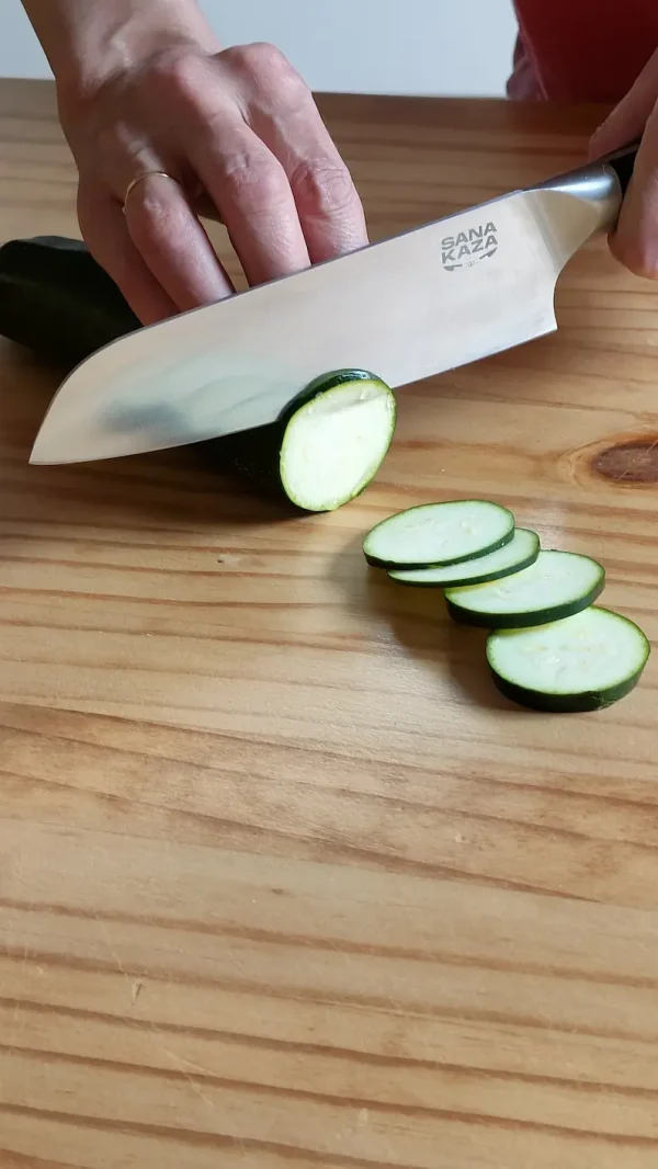 Mains utilisant un couteau de cuisine Sanakaza pour couper des rondelles fines de courgette sur une planche en bois.