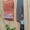 Couteau Santoku Sanakaza 18 cm à manche noir à côté d'un pavé de saumon sur une planche en bois