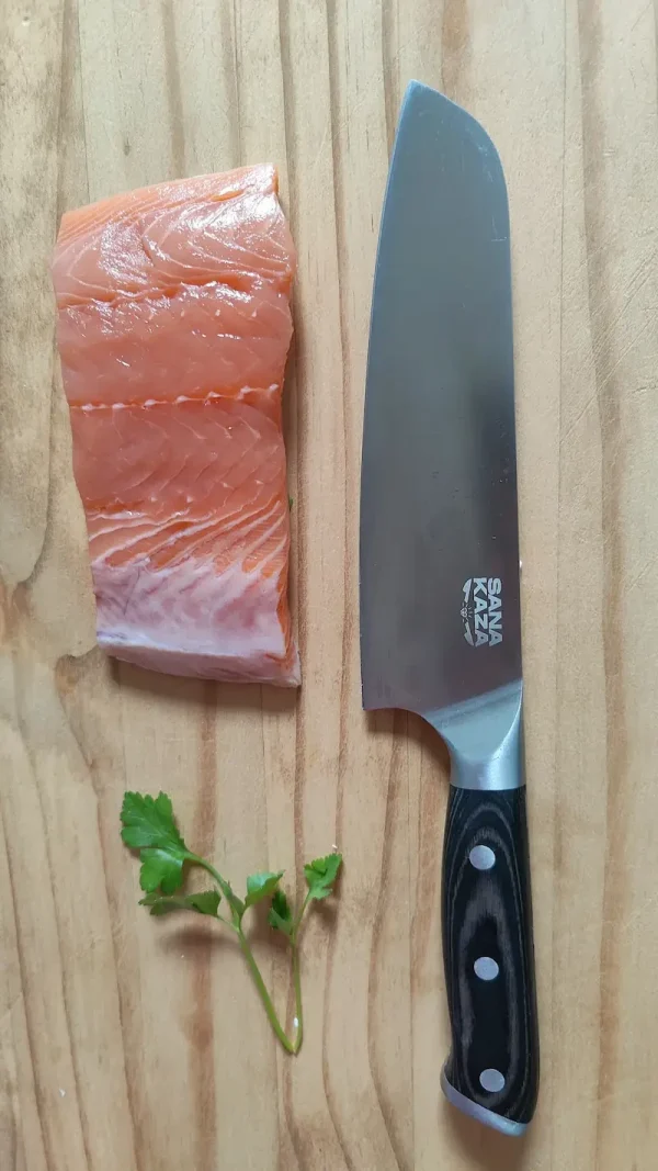 Couteau Santoku Sanakaza 18 cm à manche noir à côté d'un pavé de saumon sur une planche en bois