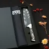 Coffret cadeau noir ouvert Sanakaza dévoilant le couteau Santoku sous son étui et le tablier noir plié sur fond sombre décoré de fleurs