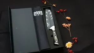 Coffret cadeau noir ouvert Sanakaza dévoilant le couteau Santoku sous son étui et le tablier noir plié sur fond sombre décoré de fleurs