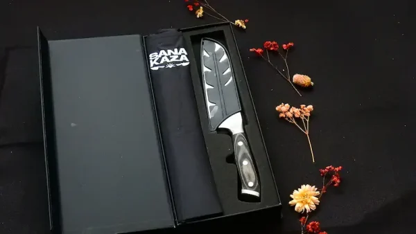 Coffret cadeau noir ouvert Sanakaza dévoilant le couteau Santoku sous son étui et le tablier noir plié sur fond sombre décoré de fleurs