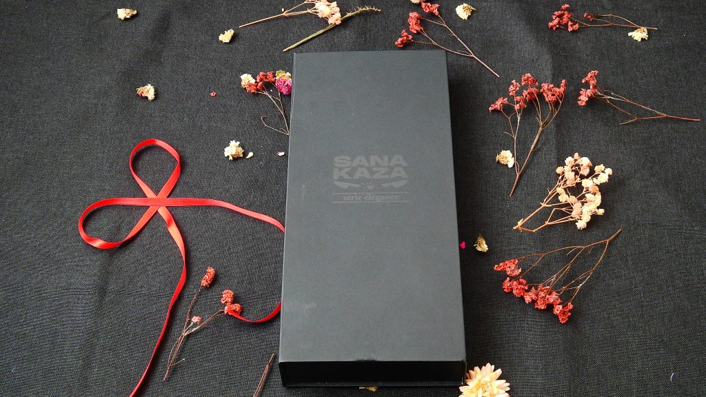 Coffret cadeau noir Sanakaza série élégance avec logo, décoré de fleurs séchées et d'un ruban rouge pour un pack couteau Santoku et tablier.