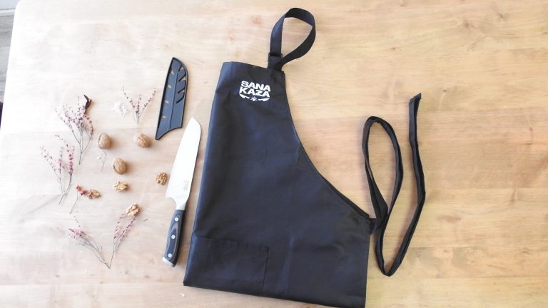 Set de cuisine Sanakaza présenté sur une table en bois : un couteau Santoku posé près de noix entières et décortiquées, avec son fourreau et un tablier noir unisexe.